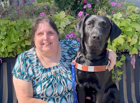 Becky and black Lab guide dog Winslow sit close on an outdoor bench for their team portrait with a garden of green and pink behind them