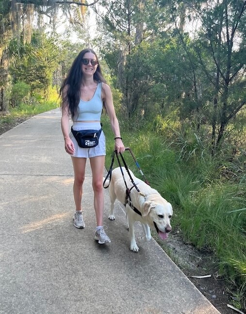 Yellow Lab Kegan leads a smiling Marin in shorts, tank and Guiding Eyes fanny pack, along a tree lined path.