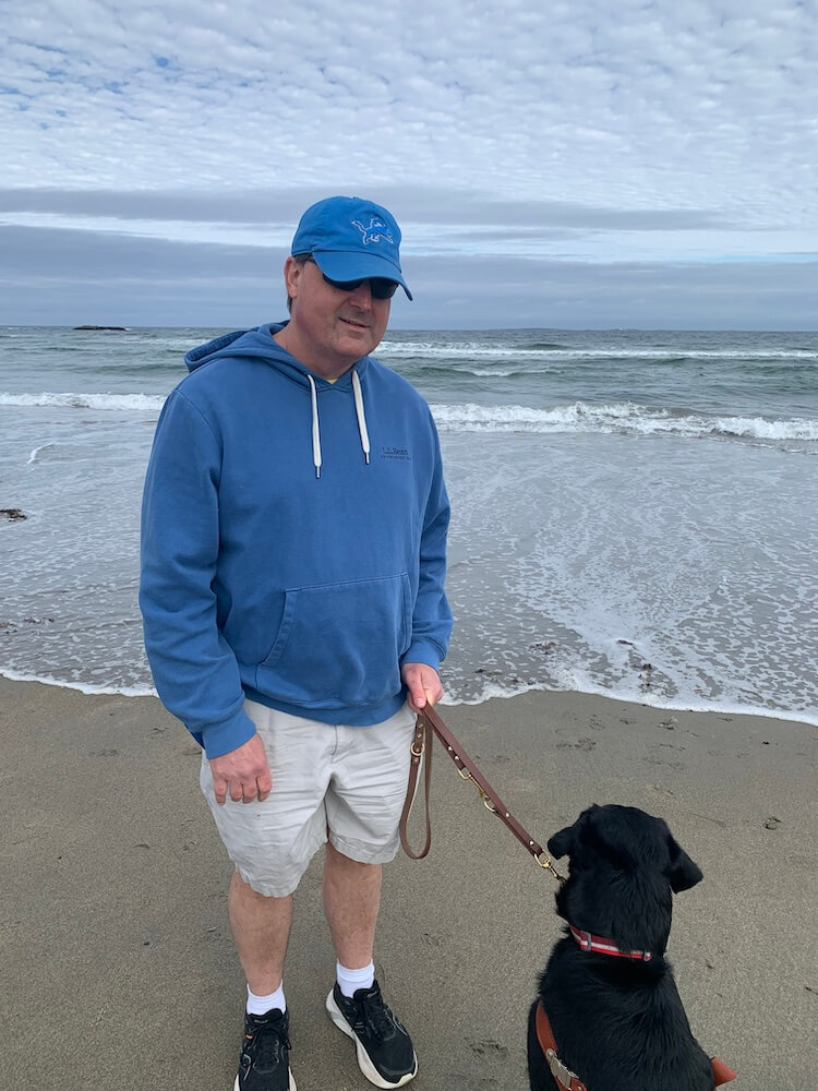 Bob and guide dog Gage stand on a wet sandy beach with waves rolling in behind them