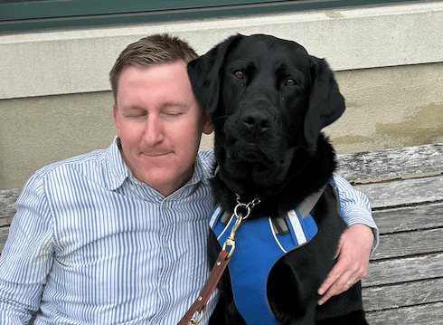 Mike leans into a hug around black Lab guide Diego sitting on an outdoor bench