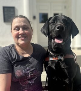 Heather and black lab guide dog Herb sit together outside for their team portrait.