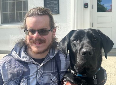 Justin sits outside on a sunny day with black guide dog Tootsie for their team portrait