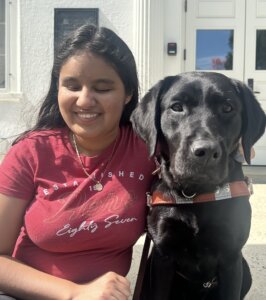 Daniela sits in a sunny area with her black Lab guide dog Sophie for their team portrait