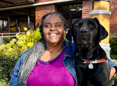 Karen and black Lab guide dog Angus sit shoulder to shoulder on a large stone area in front of a brick building for their team portrait