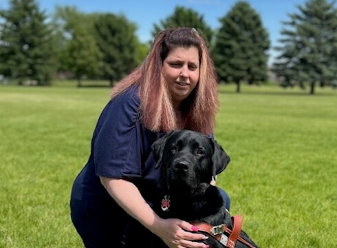 Michelle kneels on bright green lawn with hands resting on black lab guide dog Artie