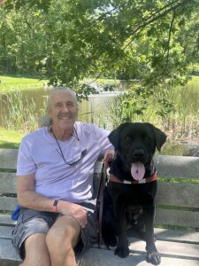 Mike and black lab guide dog Barry sit  on an outdoor bench against backdrop of greenery and pond