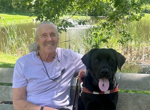 Mike and black lab guide dog Barry sit on an outdoor bench against backdrop of greenery and pond