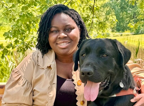 Osolu and black lab guide dog Stitch sit close on an outdoor bench against backdrop of greenery