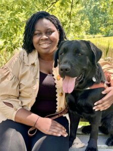 Osolu and black lab guide dog Stitch sit close on an outdoor bench against backdrop of greenery