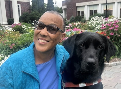 Quasi sits beside his handler Donna with her arm around him and heads together in an outdoor courtyard setting