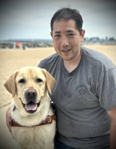 Ken kneels on the beach with an arm around yellow guide dog Tahoe