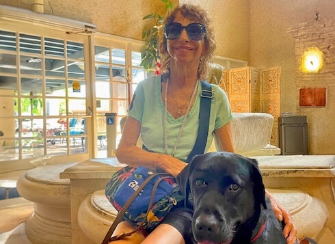 Carol and black Lab guide dog Ezra sit in a lobby with glass doors and stone benches for their team portrait