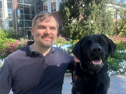 Chris and black Lab guide Wade sit against backdrop of plantings and a stately brick library