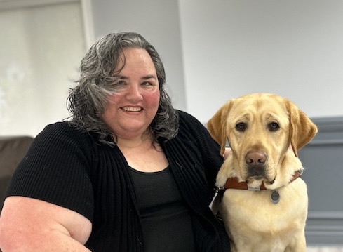 Heather and yellow Lab guide dog Darby sit together indoors for their team portrait