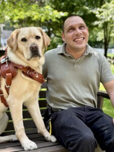 Anthony smiles happily with yellow guide dog Lindsey on  a bench in a park-like setting