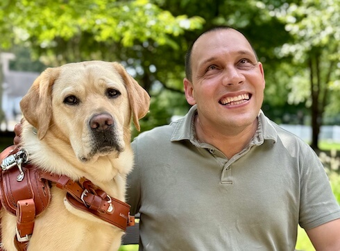 Anthony smiles happily with yellow guide dog Lindsey on a bench in a park-like setting