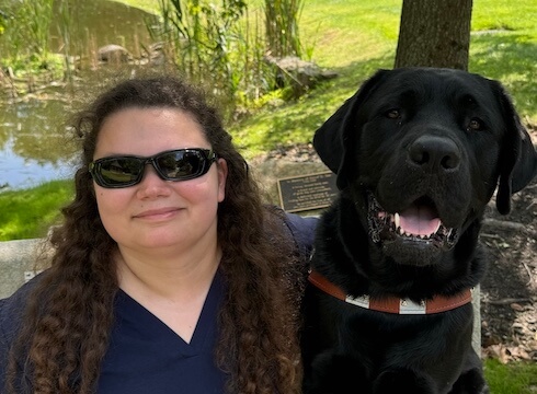 Lydia and black lab guide dog Brody sit on a bench under a tree on campus.