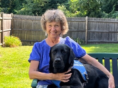 Maggee sits on an outdoor bench smiling as black lab guide dog Wegman lies across her lap with head up.