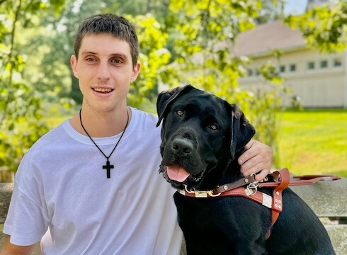 Mason and black lab guide dog Sonic sit together on an outdoor bench on campus for their team portrait