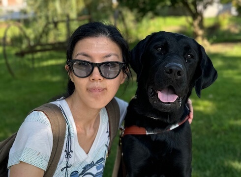 Yoojin and black lab guide Dennison sit close head to head as they sit on a bench