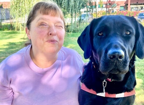 Sonia and black lab guide dog Petal sit together on an outdoor bench for team portrait