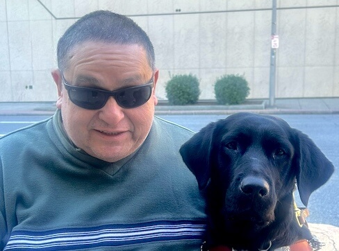 Philip and black lab guide dog Jayla sit together on an outdoor bench for team portrait