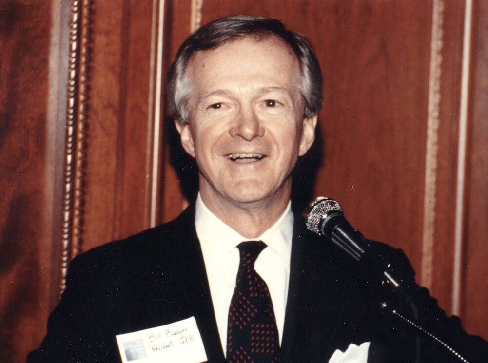 Bill Badger shown head and shoulders in a dark suit behind a podium microphone