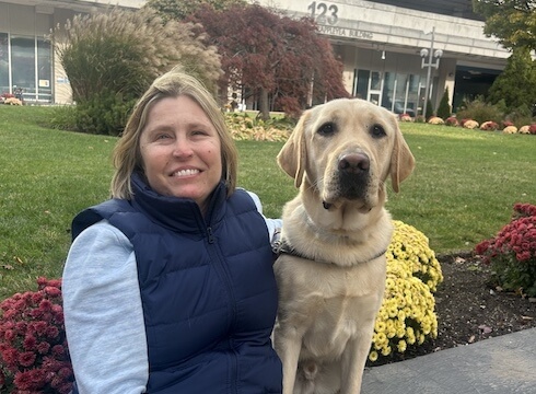 Megan sits on a retaining wall with fall mums behind her and yellow Lab guide dog Woodstock