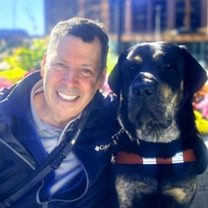 Robert and brindle lab guide dog Bingo head to head for team portrait with foliage and building in background