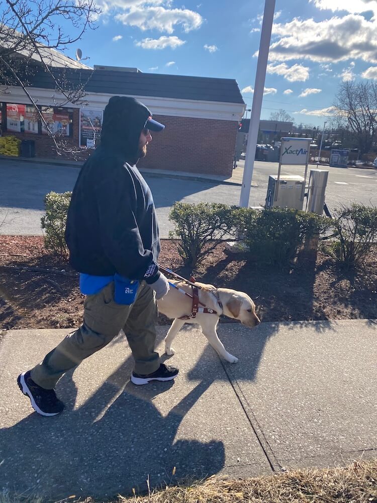 Queen leads Dustin down a sidewalk and to their left is a parking lot and convenience store