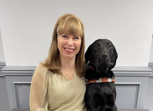 Noella sits close to black Lab guide dog Doll indoors for their team portrait