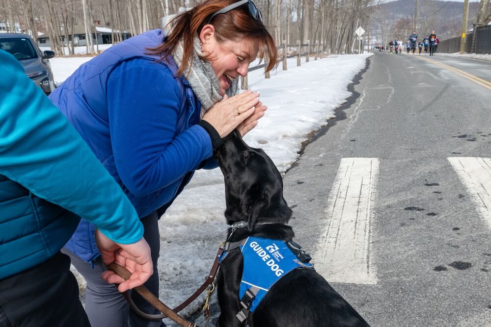 Miranda, Foundation Mgr in the Training Dept bends forward to greet Nettie, a black Lab guide dog in training wearing a Unifly harness