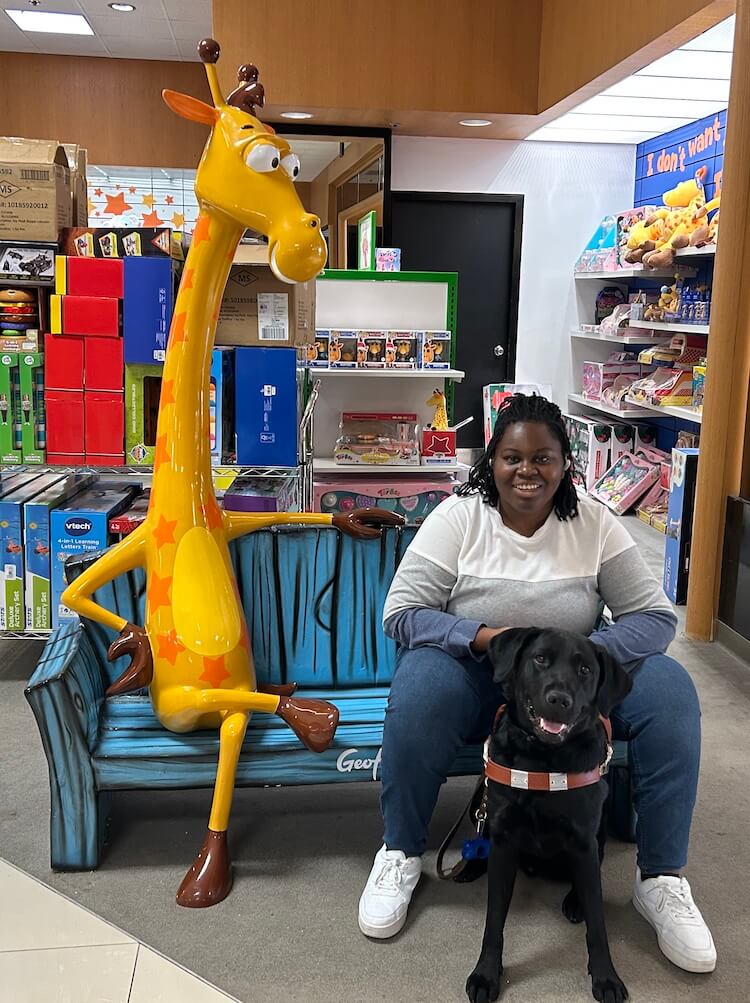 Osolu sits on a bench in a toy department with a tall bright figure of Geoffrey the  giraffe